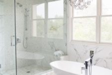 a luxurious transitional space with a fantastic chandelier, an oval tub, marble tiles and printed shades