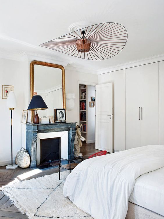 a neutral Parisian bedroom with a blue non-workign fireplace, a statement lamp, a chic mirror and an antique wooden chair