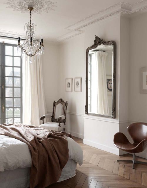 an elegant Parisian bedroom with a herringbone floor, a statement mirror, a chic crystal chandelier and a refined chair