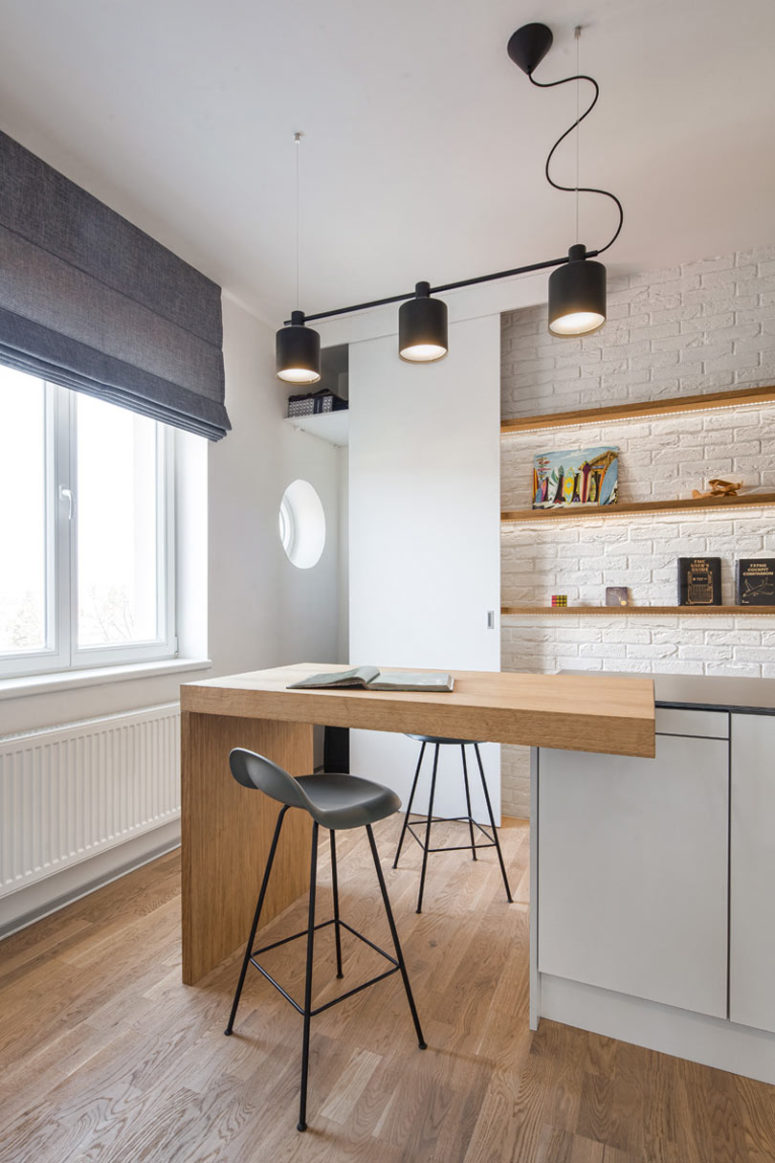 The kitchen is done with sleek minimalist cabinets, built-in shelves, a brick wall and a kitchen island that doubles as an eating space