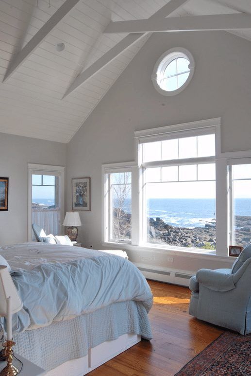 a coastal bedroom done in light blues and white, with a large window and a tiny porthole to embrace the location