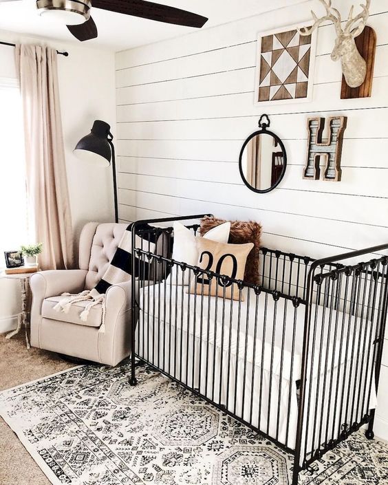 a cozy nursery with a shiplap wall, a black metal crib, a printed rug, a blush chair and blush curtains plus touches of black for drama
