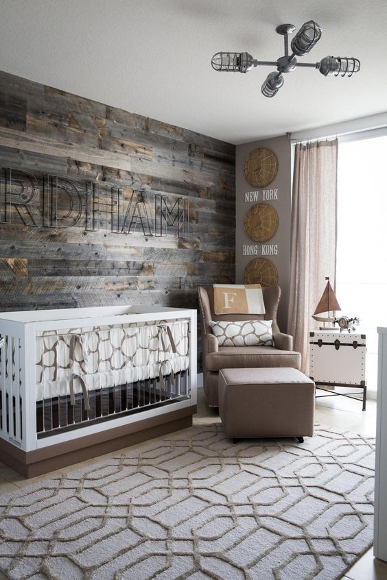 a modern farmhouse nursery with a reclaimed wooden wall, a taupe chair and footrest, a modern crib with acryl and a printed rug