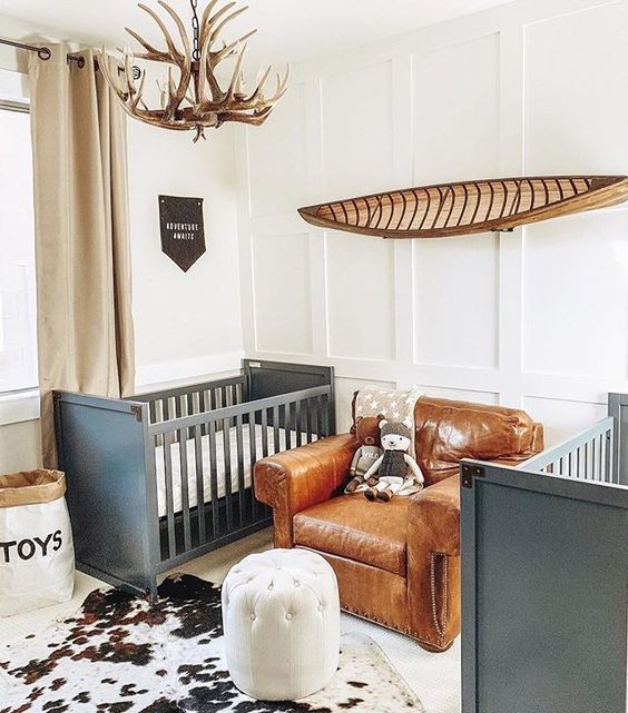 a shared farmhouse nursery with grey cribs, a leather chair, an antler chandelier and an animal skin rug