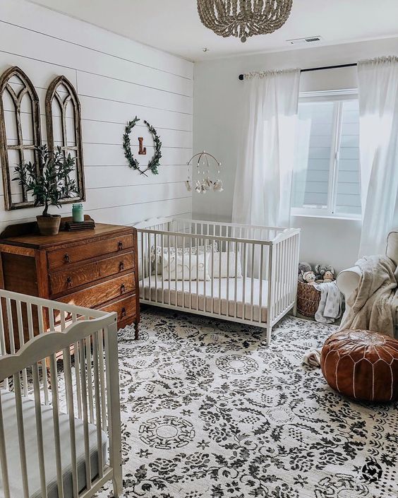 a welcoming shared farmhouse nursery with a printed rug, a leather ottoman, a stained dresser, a bead chandelier and some greenery