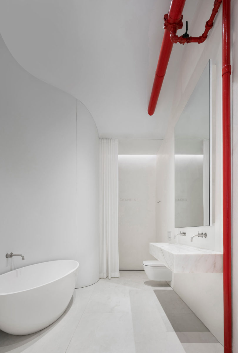 The bathroom is white and minimalist, with touches of red, an oval tub and a marble floating vanity