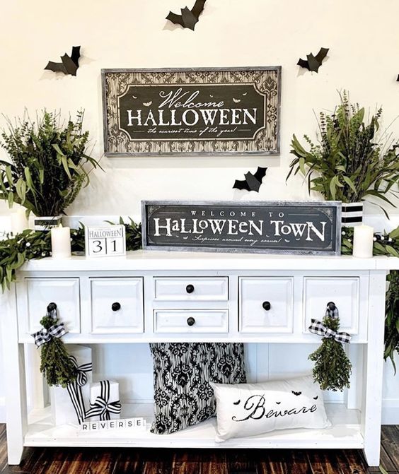 a simple and fresh Halloween entryway console with black and white pillows, greenery arrangements, signs and candles is chic