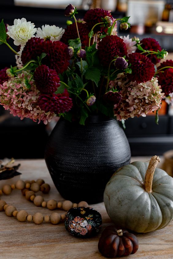 a Halloween centerpiece of a black vase, dark blooms, some pumpkins including painted ones and wooden beads