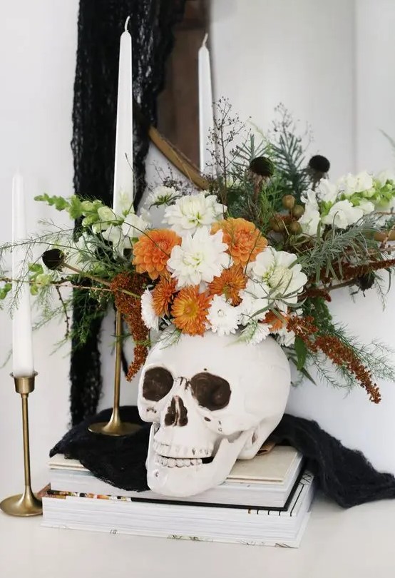 a Halloween decoration of a skull with white and orange blooms, black flowers, greenery and herbs is a nice centerpiece