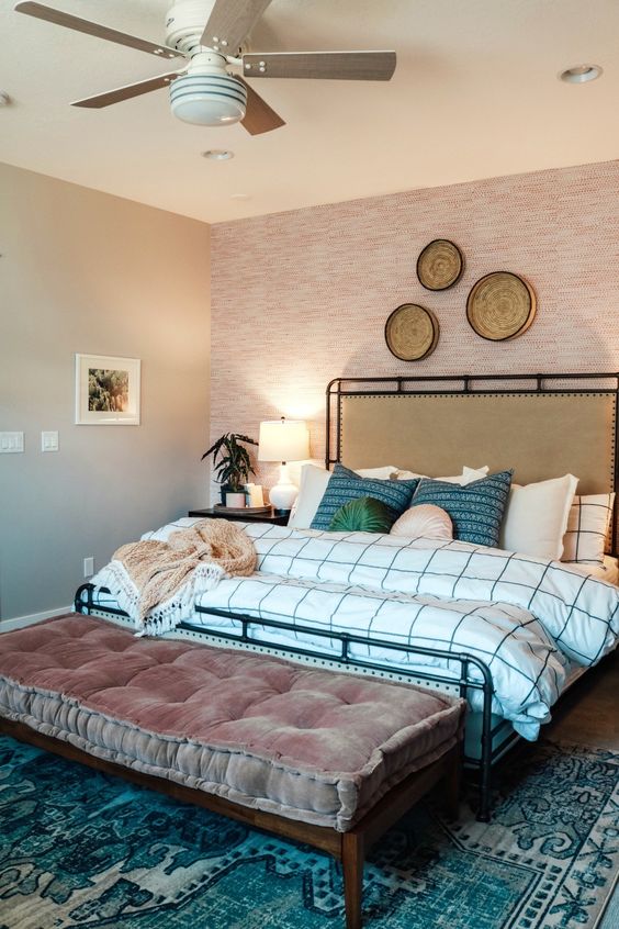 a bright boho bedroom with a pink wallpaper wall, a bed with a tan leather headboard, printed bedding, a bench with a cushion and baskets