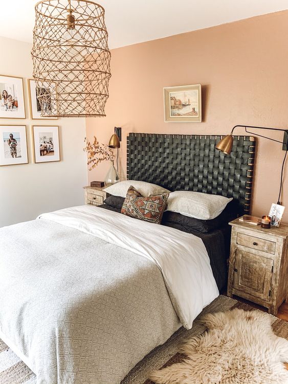 a cozy boho bedroom with a dusty pink wall, a bed with a black woven leather headboard, monochromatic bedding, brass lamps and some artwork