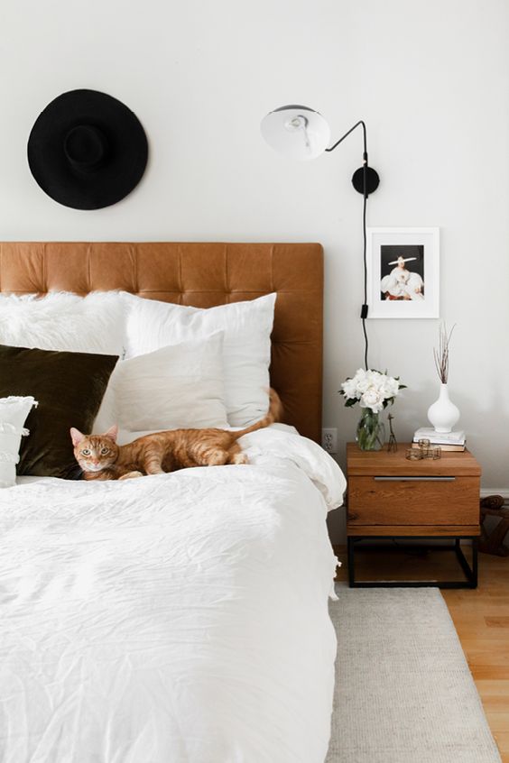 a mid-century modern bedroom with a brown leather bed, monochromatic bedding, stained nightstands, a sconce and some decor