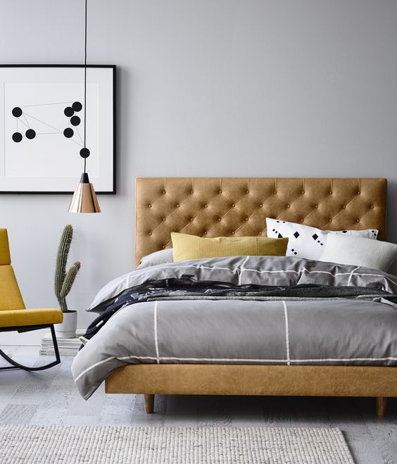a modern bedroom with an amber leather bed, grey bedding, a yellow rocker chair, a copper pendant lamp
