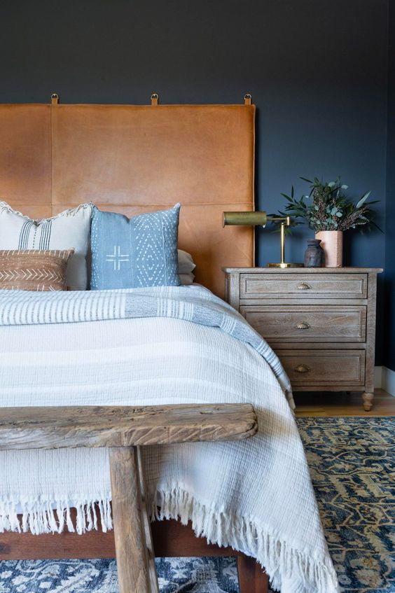 a modern boho bedroom with navy walls, a bed with an amber leather headboard, a reclaimed wood bench and nightstand