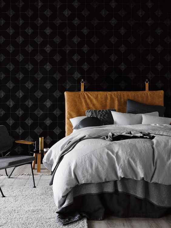a moody bedroom with black wallpaper, a bed with monochromatic bedding, an amber leather hanging headboard, a nightstand and a grey chair