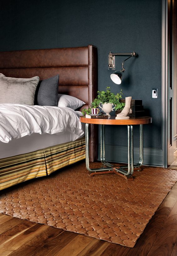a moody bedroom with soot walls, a bed with a brown leather headboard, a nightstand with greenery and a brown rug