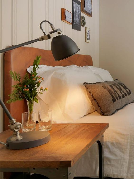 a rustic bedroom with an amber leather bed, white bedding, a reclaimed wood nightstand, a black lamp and a gallery wall