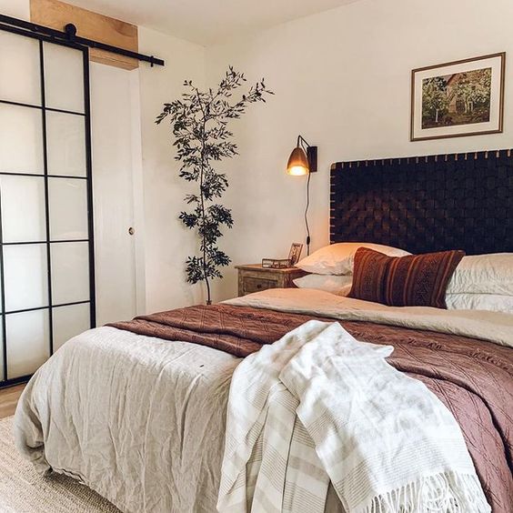 a stylish bedroom with a black woven headboard, neutral bedding, a stained nightstand, a sconce and greenery
