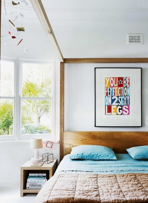 a white bedroom with natural wooden furniture is made brighter with blue bedding and a colorful artwork on the wall