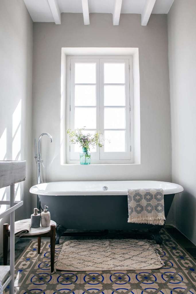 The bathroom is rustic, with a vintage tub, tiles, wooden beams and a large window