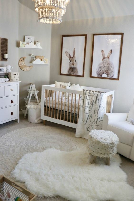 a cozy neutral nursery with artworks, a crib, layered rugs, a fringe stool and some simple wooden furniture