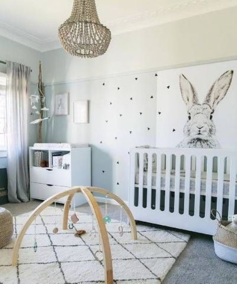 a fun neutral nursery with a creative wall, white modern furniture, a printed rug, a beaded chandelier and baskets for storage