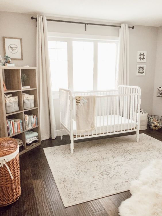a light-filled neutral nursery with a crib, a wooden shelving unit, a printed rug, baskets and neutral curtains