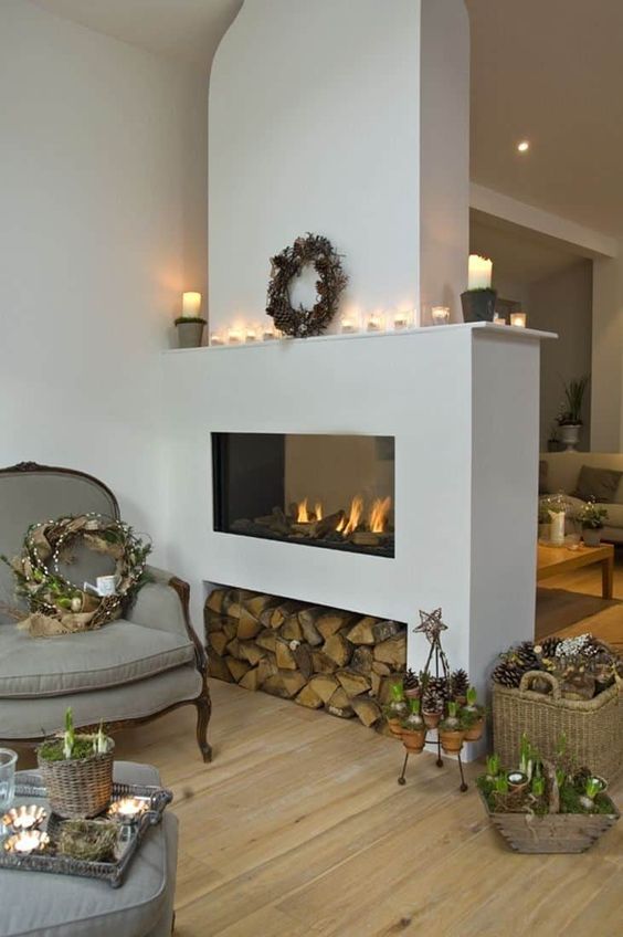 a sleek white fireplace with firewood storage, candles on the mantel and a wreath with pinecones for winter