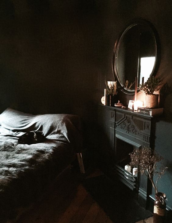 a gorgeous dark moody bedroom with a faux fireplace, a dark framed mirror and a bed with black bedding