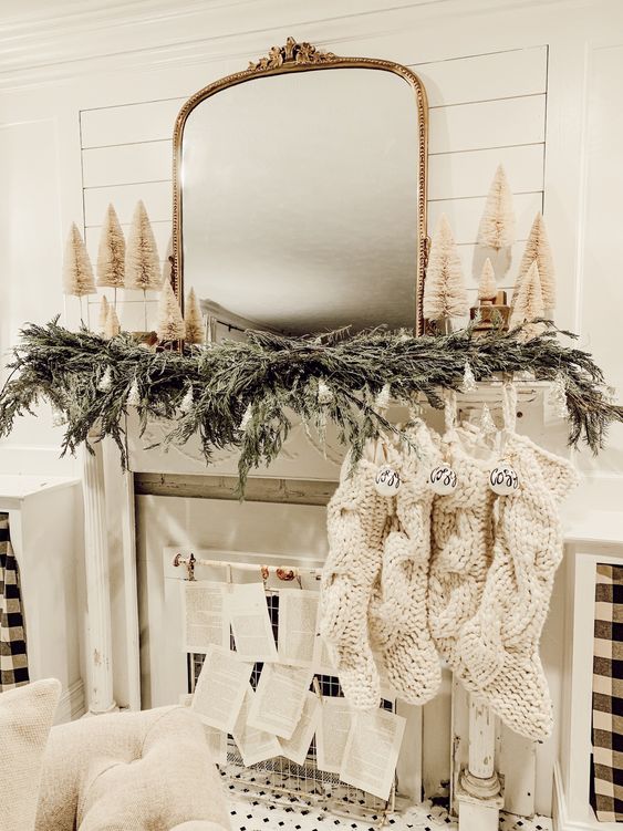 a cozy neutral winter fireplace with an evergreen garland, mini bottle bruch trees, knit stockings with ornaments and book pages