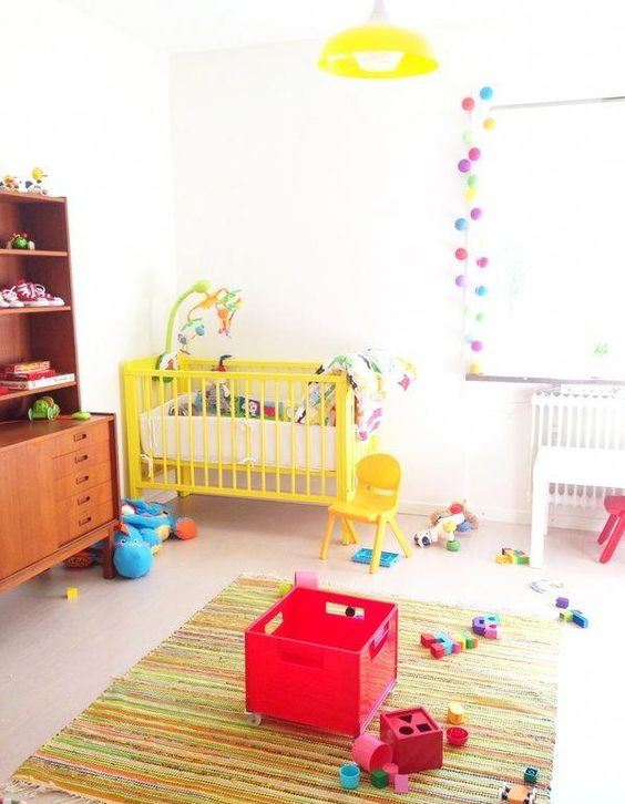 a bright and fun nursery with a printed rug, a yellow baby crib, colorful pompom garlands and bright toys
