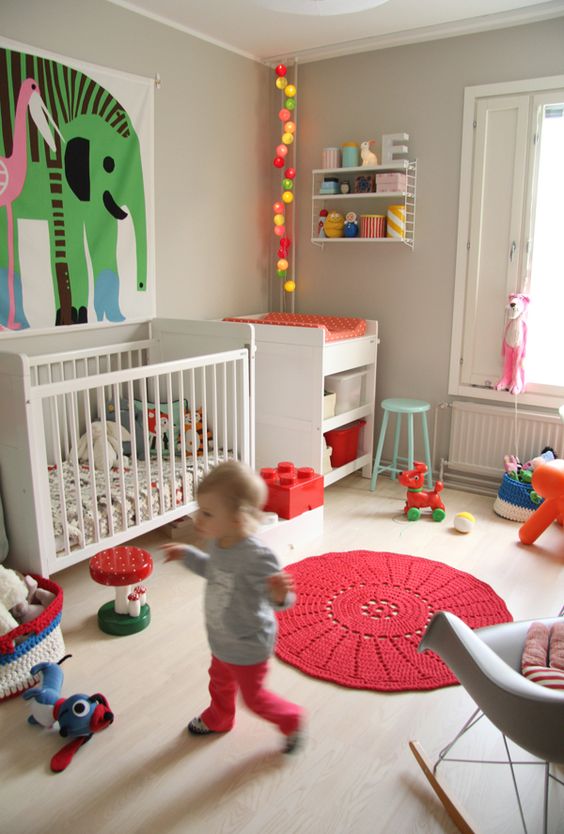 a bright and fun nursery with a whimsy artwork, colorful light garland, colorful rugs, toys and crochet baskets