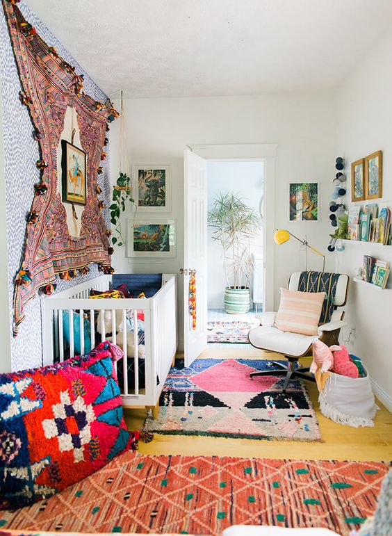 a bright boho nursery with a pompom rug on the wall, colorful rugs on the floor, pillows and bright bedding