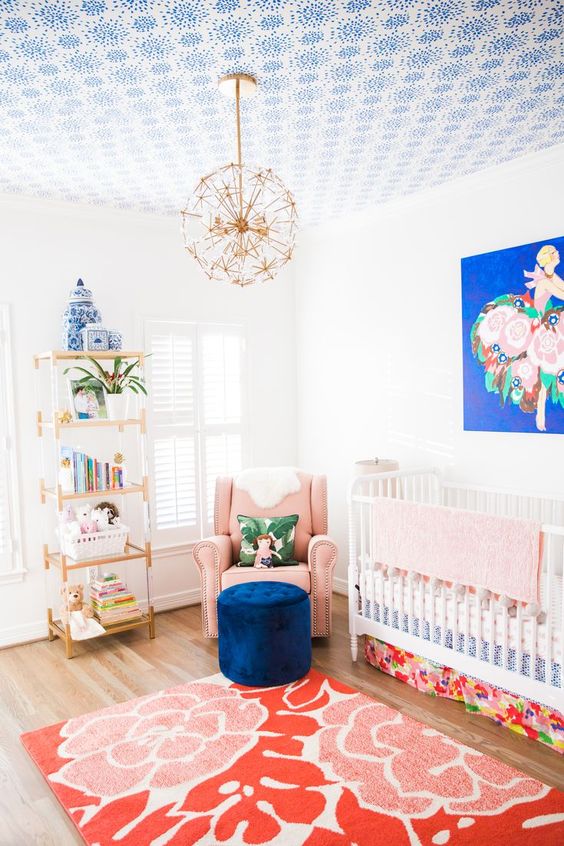 a brigth mixed pattern nursery with a floral rug, a navy ottoman, a brigth artwork and colorful books