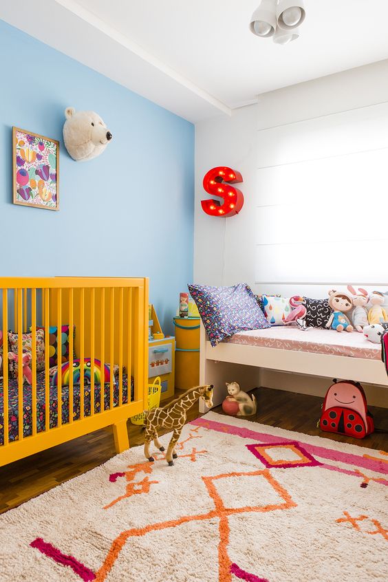 a brigth nrusery with a yellow crib, a printed rug, a marquee letter, bright toys and artworks for fun