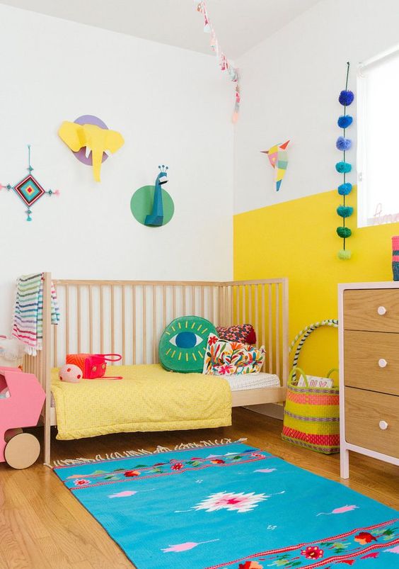 a colorful nursery with a color block wall, a printed rug, colorful mobiles and fun art on the wall plus brigth bedding