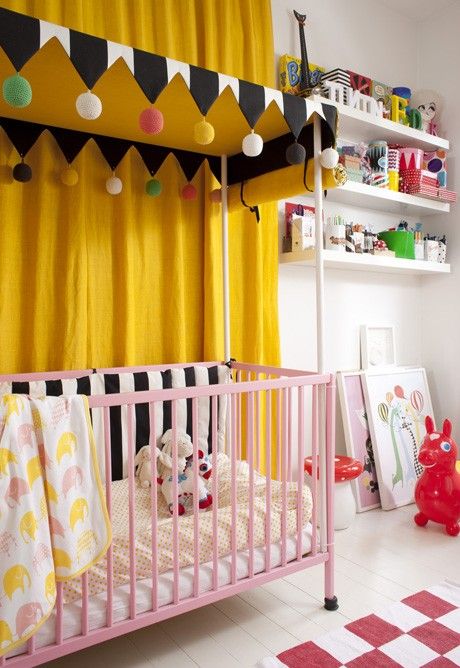 a colorful nursery with yellow curtains, a pink crib, a printed rug, colorful accessories and lots of toys