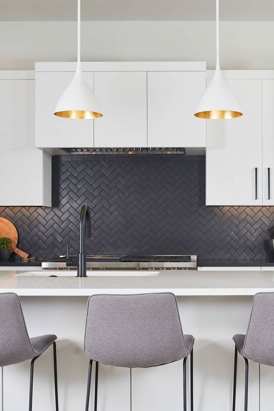 a contemporary white kithen with a black herringbone backsplash, tall grey stools and pendant lamps with gold inner