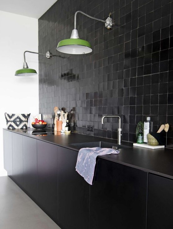 a cool black kitchen with sleek lower cabinets, tiles all over the wall, green lamps and stone countertops