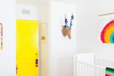 a super colorful nursery with a bright yellow door, colorful pompoms and tassels, a rug, a bright ottoman and bedding