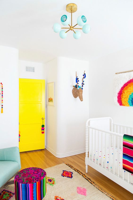 a super colorful nursery with a bright yellow door, colorful pompoms and tassels, a rug, a bright ottoman and bedding