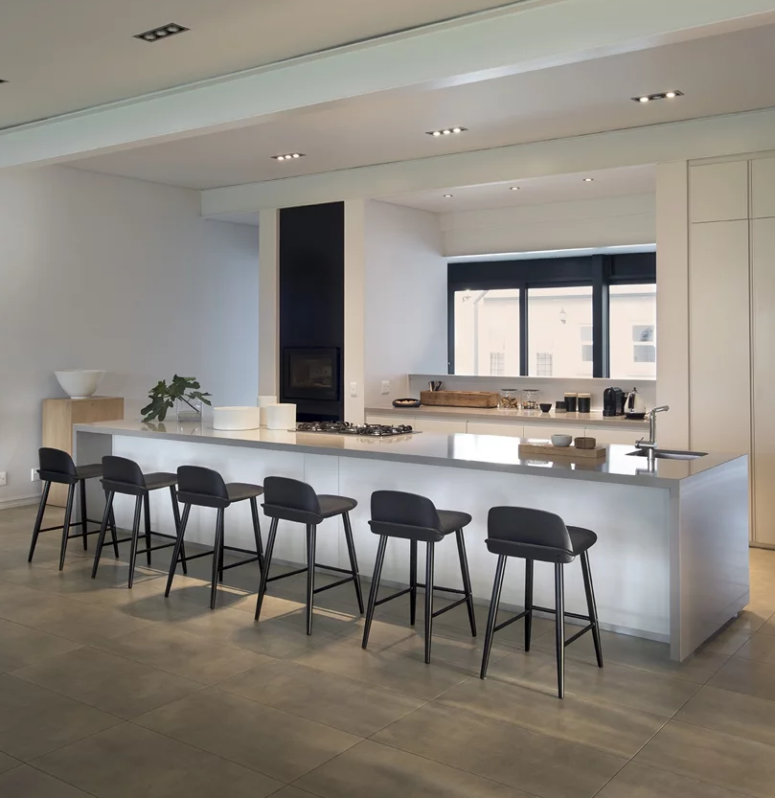 The kitchen is all white and sleek, with a giant kitchen island, a window to bring light in and black stools The kitchen is all-white and sleek, with a giant kitchen island, a window to bring light in and black stools