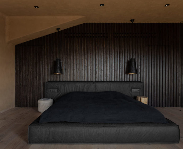 The bedroom is dark and soothing, with a wood clad wall, an upholstered dark bed and wall sconces