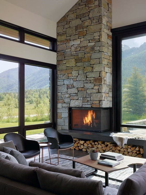 a contemporary cabin living room with a stone clad fireplace and firewood storage, chic furniture and lots of windows