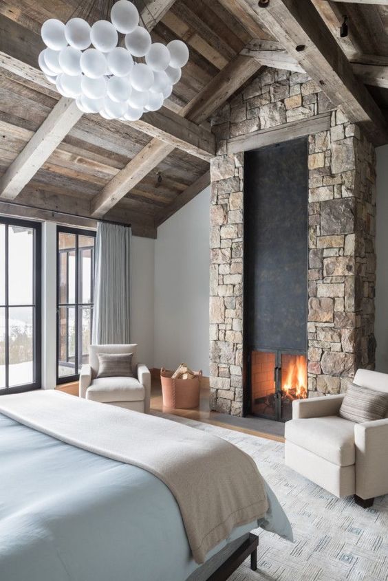 a modern chalet bedroom with a wooden ceiling, a bed with blue bedding, a stone clad fireplace and a bubble chandelier