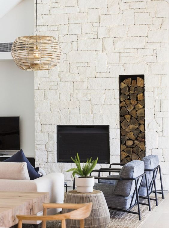 a modern coastal living room with a stone fireplace, a navy sofa, grey chairs, a side table and a woven pendant lamp