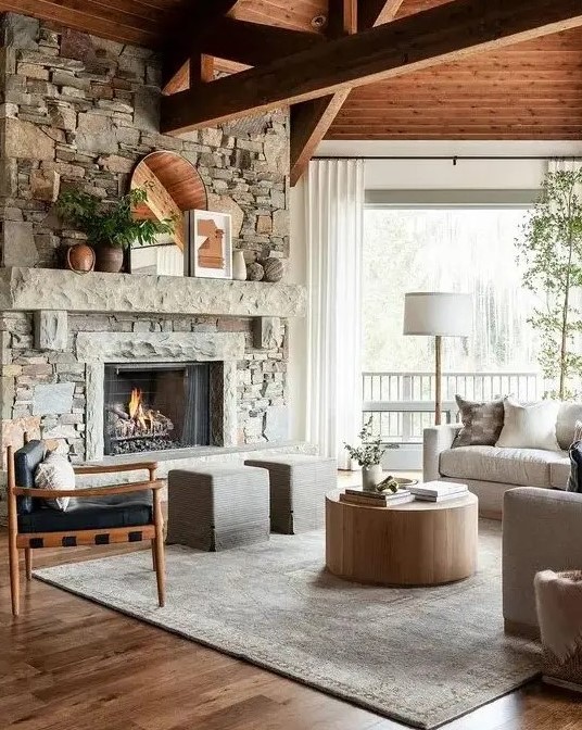 a modern country living room with a fireplace clad with faux stone, wooden beams, neutral furniture and a round coffee table