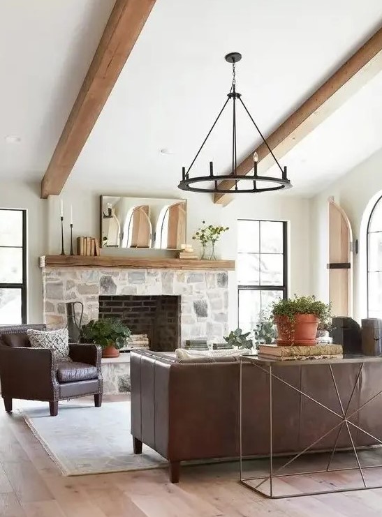 a modern farmhouse living room with a stone clad fireplace, wooden beams, leather furniture, a metal chandelier and potted plants