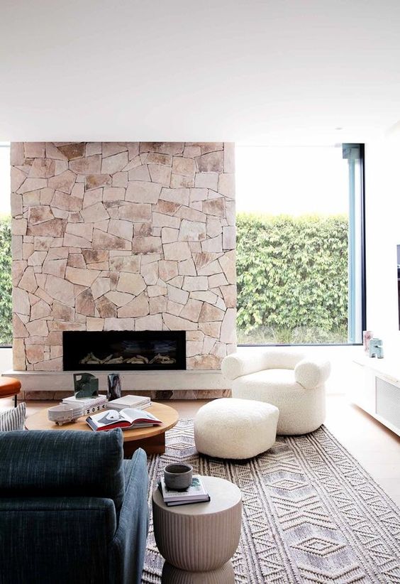 a modern living room with a stone fireplace, an indigo sofa, low coffee tables and a white chair and pouf is wow
