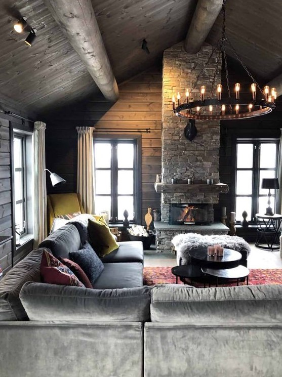 a moody chalet living room with wooden walls and a ceiling, a stone fireplace, a chandelier and comfortable and stylish furniture